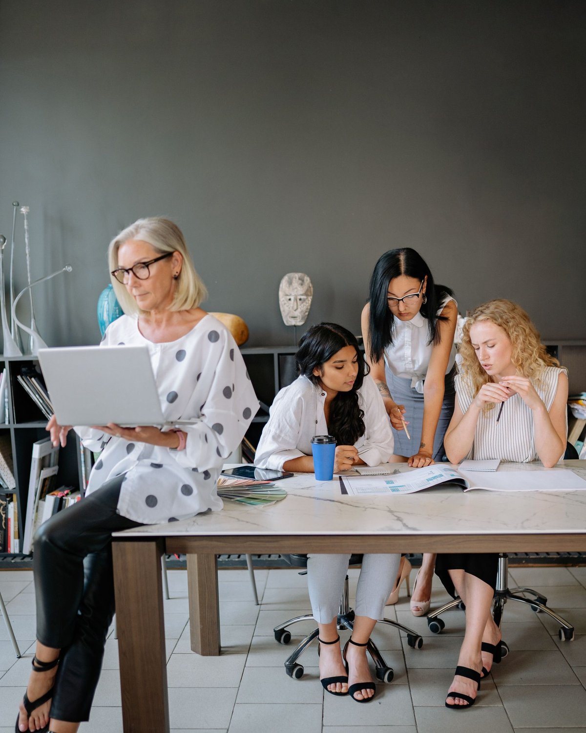 A mixed age team of women working together in an office, an experienced colleague with a laptop alongside younger colleagues around a table